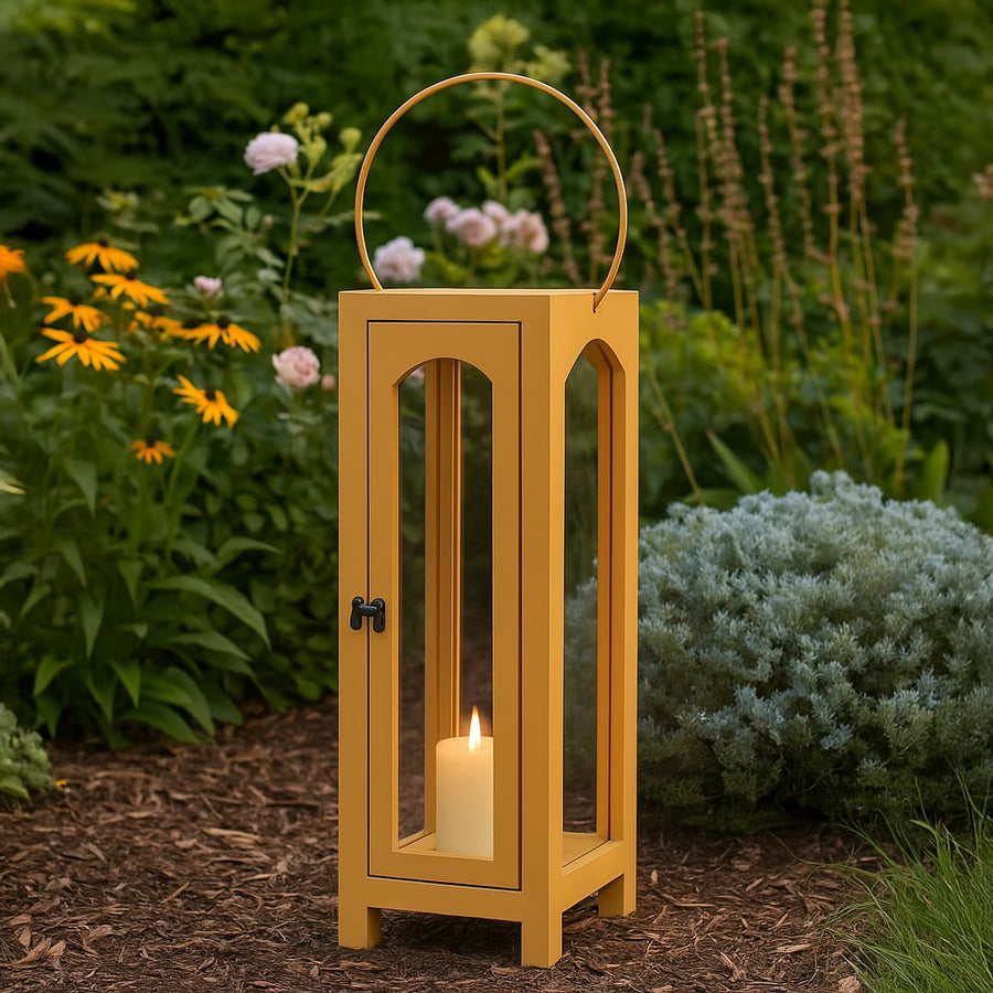Natural wood floor lantern with a lit candle placed in a garden bed surrounded by plants creating a cozy warm outdoor ambiance.