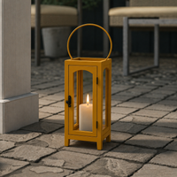 Modern mustard yellow tall frame glass lantern floor candle holder standing on a stone tiled outdoor patio courtyard floor.