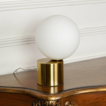 Contemporary round white glass table lamp with gold metal base placed on wooden table against minimal wall background perfect for ambient lighting.