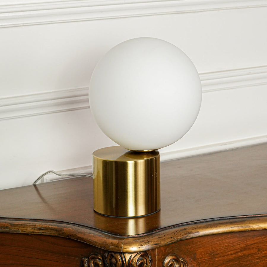 Contemporary round white glass table lamp with gold metal base placed on wooden table against minimal wall background perfect for ambient lighting.
