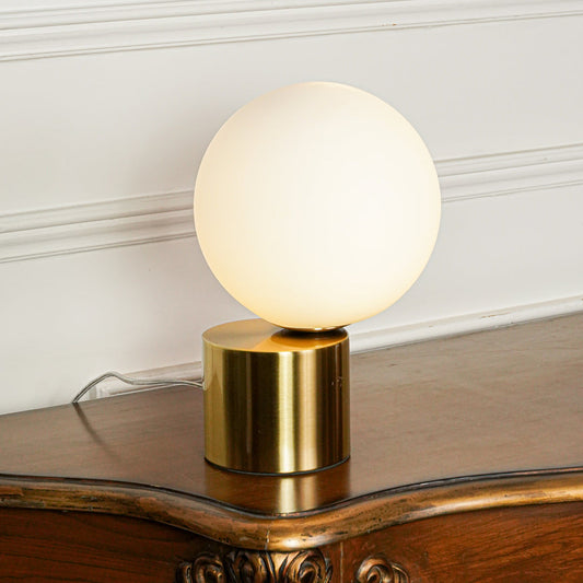 Elegant frosted glass globe table lamp with brass finish base set on wooden surface with plant decor and neutral wall background for modern interiors.