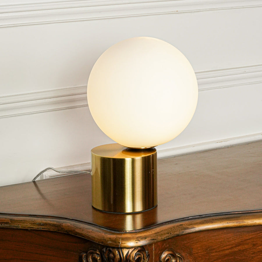 Elegant frosted glass globe table lamp with brass finish base set on wooden surface with plant decor and neutral wall background for modern interiors.