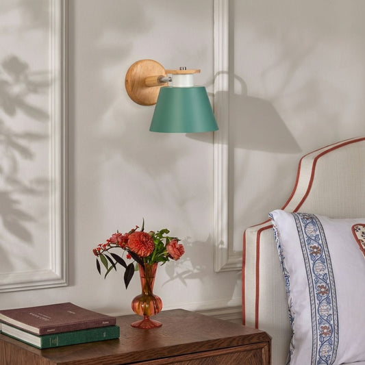 A wall-mounted sconce with a teal-green conical shade, a light wood arm, and a round wooden backplate, shown lit in a bedroom setting next to a bed and a small vase of flowers.