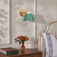 A wall-mounted sconce with a teal-green conical shade, a light wood arm, and a round wooden backplate, shown lit in a bedroom setting next to a bed and a small vase of flowers.