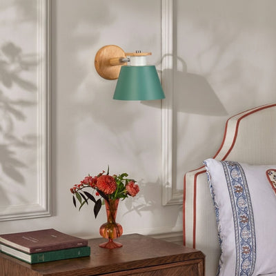A wall-mounted sconce with a teal-green conical shade, a light wood arm, and a round wooden backplate, shown lit in a bedroom setting next to a bed and a small vase of flowers.