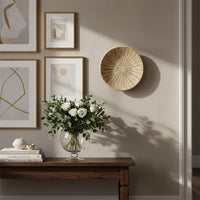 A beige woven bowl hangs on a wall next to framed art above a wooden table holding a glass vase with white roses and books in sunlight.