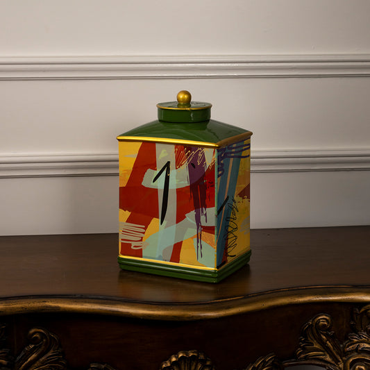 Decorative ceramic jar with colorful abstract pattern and green lid placed on wooden table against neutral wall background showcasing vibrant artistic home decor styling.