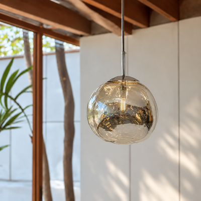 A modern spherical pendant light with a textured, hammered clear glass shade and a polished silver-toned metal rod, hanging in a contemporary indoor setting with warm wood ceiling beams and soft natural light.