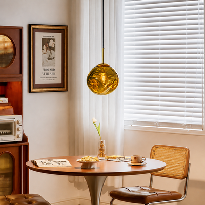 A round wooden dining table in a cozy room setting, styled with a brown leather and cane chair, a hanging amber glass pendant light, and a framed print on the wall.