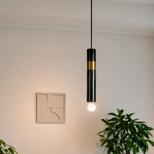 A modern black pendant light with a polished gold accent band and a glowing light bulb, hanging in a minimalist room with a beige wall, abstract art, and a green plant.
