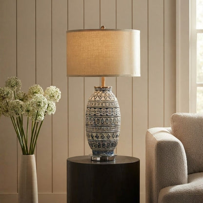 A table lamp with a textured beige drum shade and a blue-and-white patterned ceramic base, styled on a dark round side table in a living room setting next to a chair and a vase of white flowers.