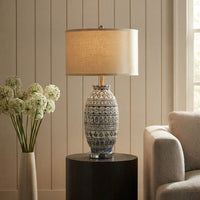 A table lamp with a textured beige drum shade and a blue-and-white patterned ceramic base, styled on a dark round side table in a living room setting next to a chair and a vase of white flowers.