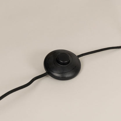Close up of a round black foot switch attached to a gold floor lamp power cord resting on a light surface.