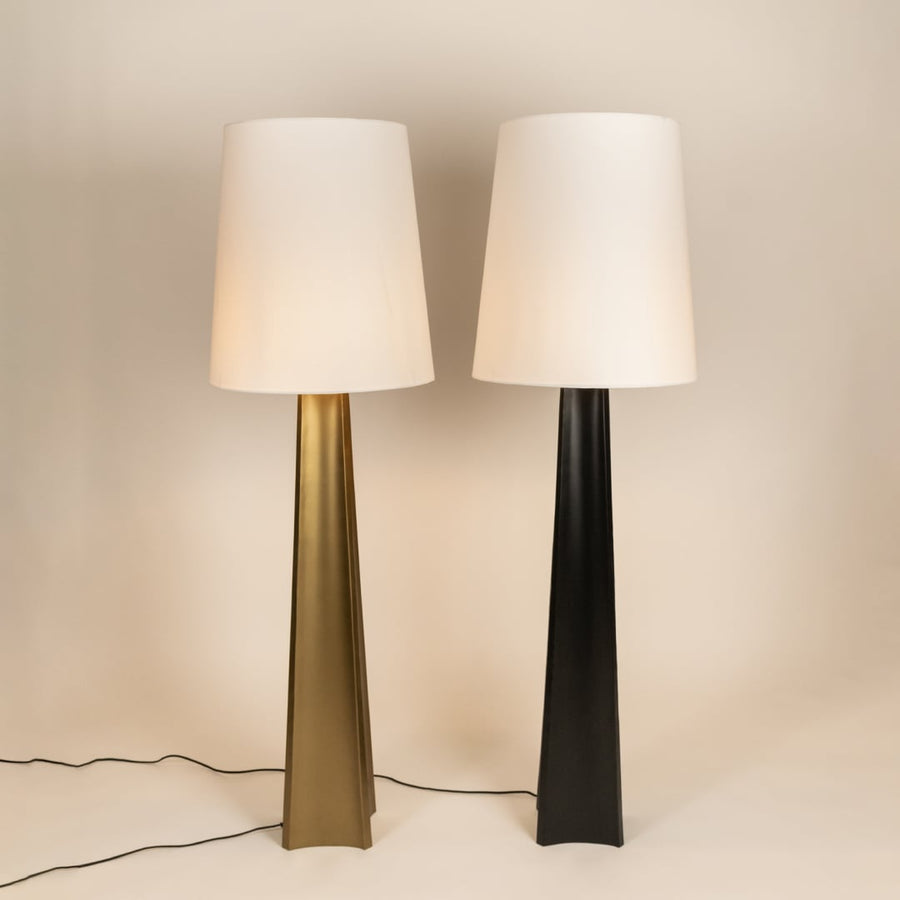 Two tall floor lamps with white fabric shades and sleek tapered bases in gold and black placed on a light surface with visible power cords.