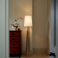 Brass floor lamp with white fabric shade casting warm ambient light in a cozy hallway and bedroom interior setting.