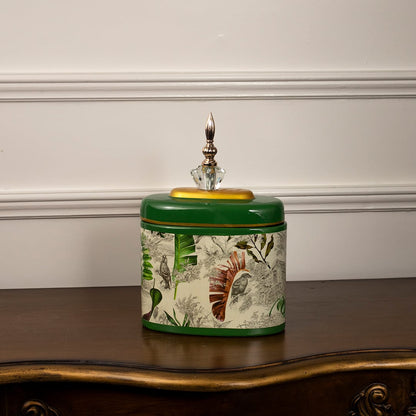 Green tropical jar with golden lid and crystal finial featuring detailed tropical leaves on white background.