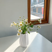 A white ribbed ceramic vase filled with white daisies and green stems, placed on a white table next to a wooden window frame.