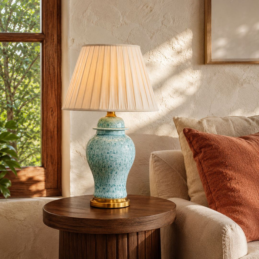 A table lamp with a speckled light blue ceramic base and a cream pleated shade, lit and placed on a round wooden side table next to a beige sofa with a rust-colored pillow in a sunlit room.