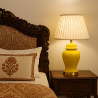Yellow ceramic table lamp with golden base and pleated fabric shade styled on wooden bedside table.
