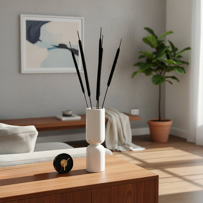 Minimal white ceramic vase with tall black decorative stems placed on wooden console beside clock sofa and plant creating calm modern living room style.
