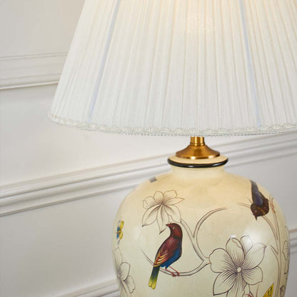 Closeup of ceramic lamp base featuring colorful bird and floral artwork on ivory finish with golden base detailing.