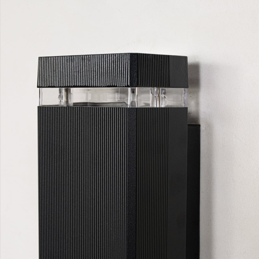 Closeup view of black metal wall light showing ribbed surface finish and clear glass diffuser for premium modern lighting design.