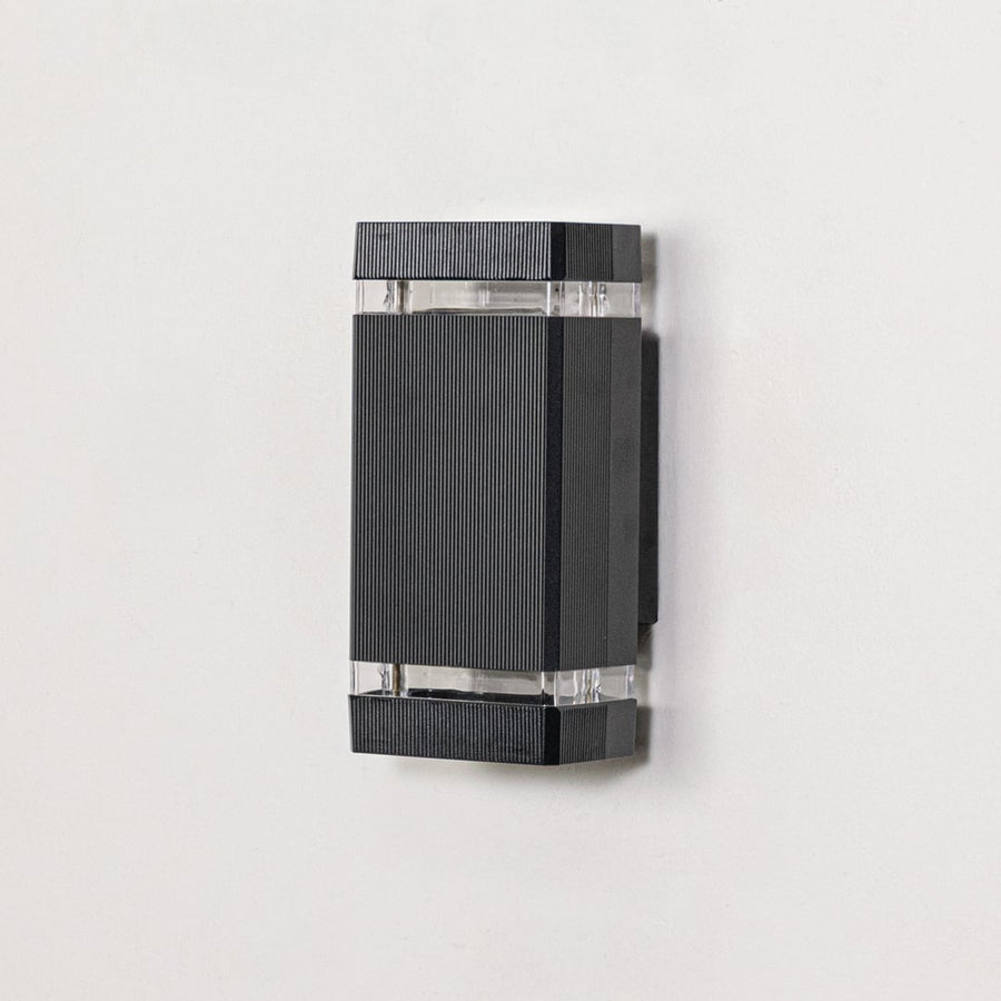 Rectangular black metal wall mounted light with ribbed finish and clear glass section displayed off on clean white background.