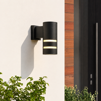 Modern black aluminum cylindrical grid downlight wall sconce on white exterior building facade next to front entryway door.