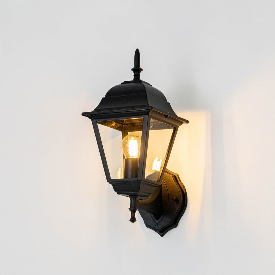 Outdoor wall light in black metal illuminated on white background radiating soft yellow glow through clear glass panes.