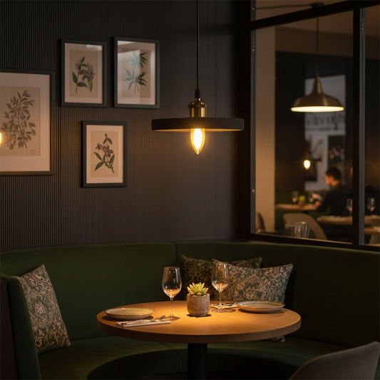 Black ring pendant light glowing warmly over a round wooden dining table with elegant green seating in a cozy restaurant setting.