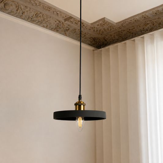 A modern black circular pendant light with a brass fixture and a clear glass bulb, hanging from a black cord in an interior room with cream walls and ornate molding.