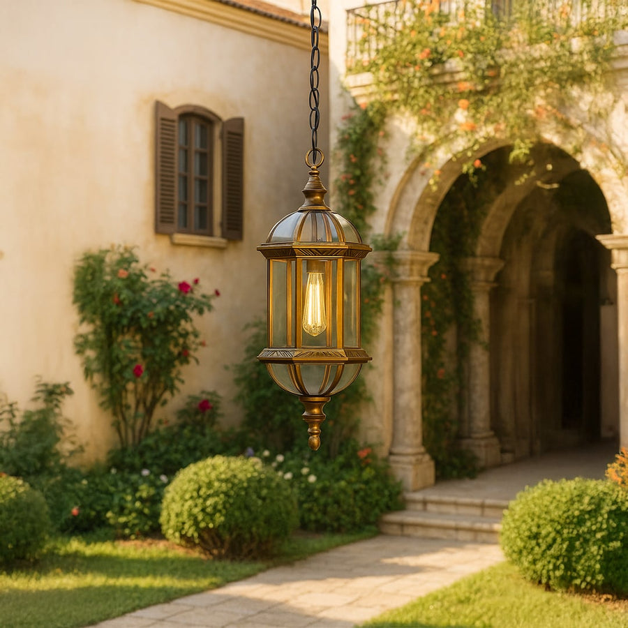 Antique brass outdoor pendant light hanging in a garden archway with warm filament bulb glowing against a blurred stone background.