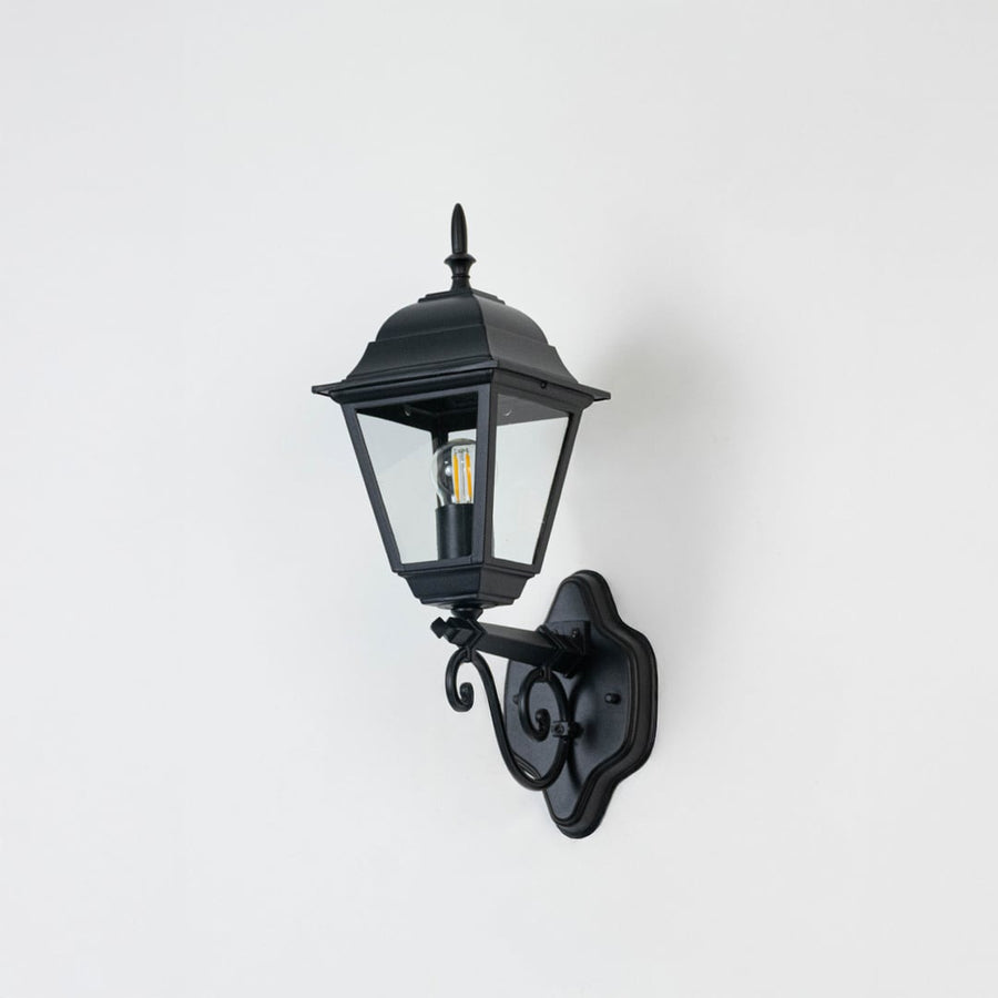 Elegant black outdoor wall lantern unlit on white background showing detailed metal frame and clear glass body in focus.