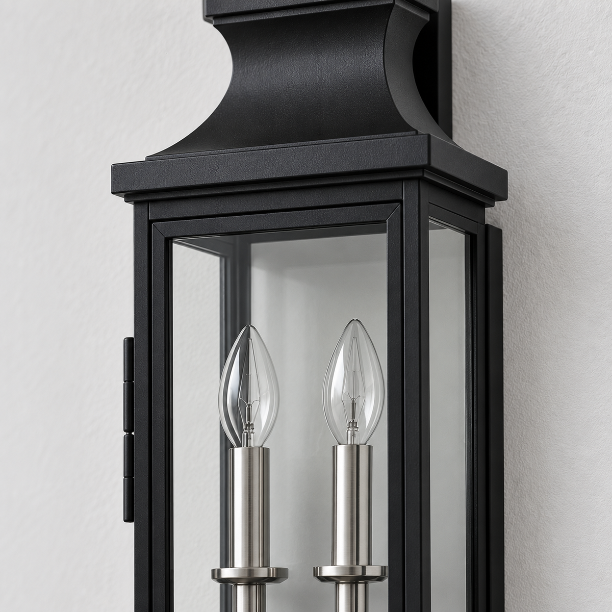 Black lantern-style wall sconce with two clear glass candle bulbs and silver metal accents, shown mounted against a light textured wall.