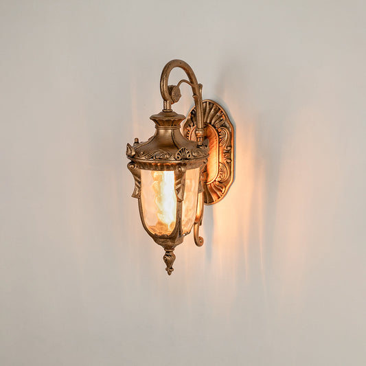 Antique brass metal outdoor wall light on white background with ornate scroll accents and clear glass shade for decor.