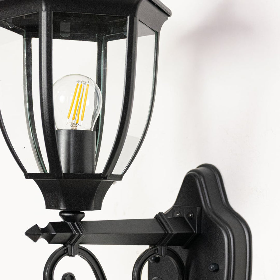 Closeup of Victorian style outdoor wall light facing upward in black metal showing decorative curves glass detail and classic exterior charm.