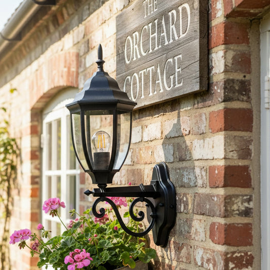 Outdoor wall lantern mounted on brick exterior beside cottage entrance creating warm welcoming appeal with classic black metal design and garden flowers accent.