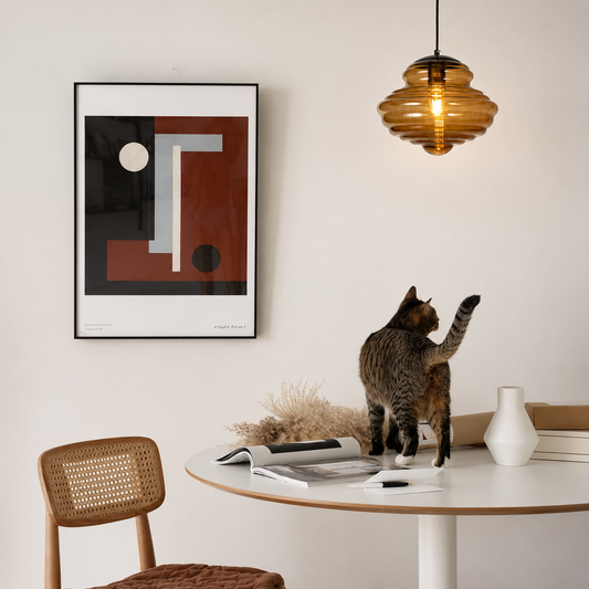 A modern interior scene with a cat standing on a round white table, looking at a framed abstract art print on the wall. The room includes a warm amber glass pendant light, a woven-back chair, a white vase, and a cat on the tabletop.