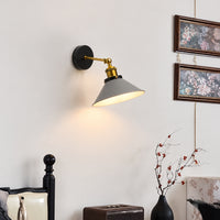 A wall-mounted swing-arm sconce lamp with a matte black round backplate, gold-toned metal arm, and a gray conical shade, shown lit on a white wall in a styled interior setting.