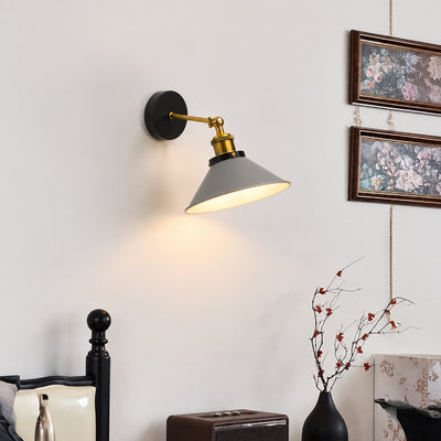 A wall-mounted swing-arm sconce lamp with a matte black round backplate, gold-toned metal arm, and a gray conical shade, shown lit on a white wall in a styled interior setting.