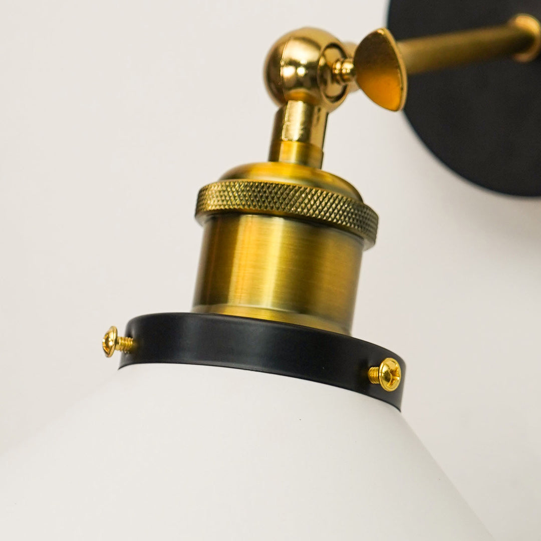 Close-up of a wall-mounted lamp with a white glass shade, brass-finished metal fixture, and a black mounting band with gold screws against a white wall.