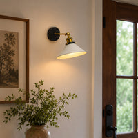 A wall-mounted sconce with a white conical shade, brass-toned hardware, and a black round backplate, lit and mounted on a beige wall next to a wooden door frame.
