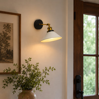 A wall-mounted sconce with a white conical shade, brass-toned hardware, and a black round backplate, lit and mounted on a beige wall next to a wooden door frame.