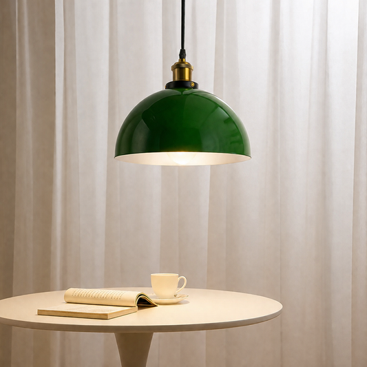 A green dome pendant light hangs over a small round table with an open book and a cup of coffee, set against a soft beige curtain background.