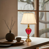Table lamp with a red base and gold accents on a table next to a vase and small bowl, with a window in the background.