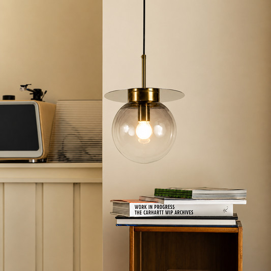 A modern pendant light with a clear glass globe shade and a brass-finished rod and canopy, hanging in a warm beige room above a small wooden side table with stacked books.