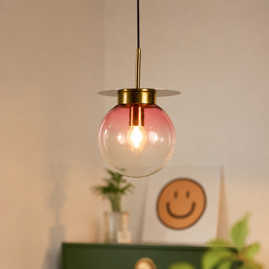 A modern pendant light with a round pink-tinted glass globe shade, a brass-colored ceiling mount, and a black hanging cord, shown in a softly lit interior setting.