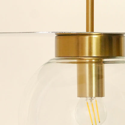 Close-up of a hanging light fixture with a brushed brass metal body, a clear glass shade, and an exposed filament bulb against a warm off-white background.