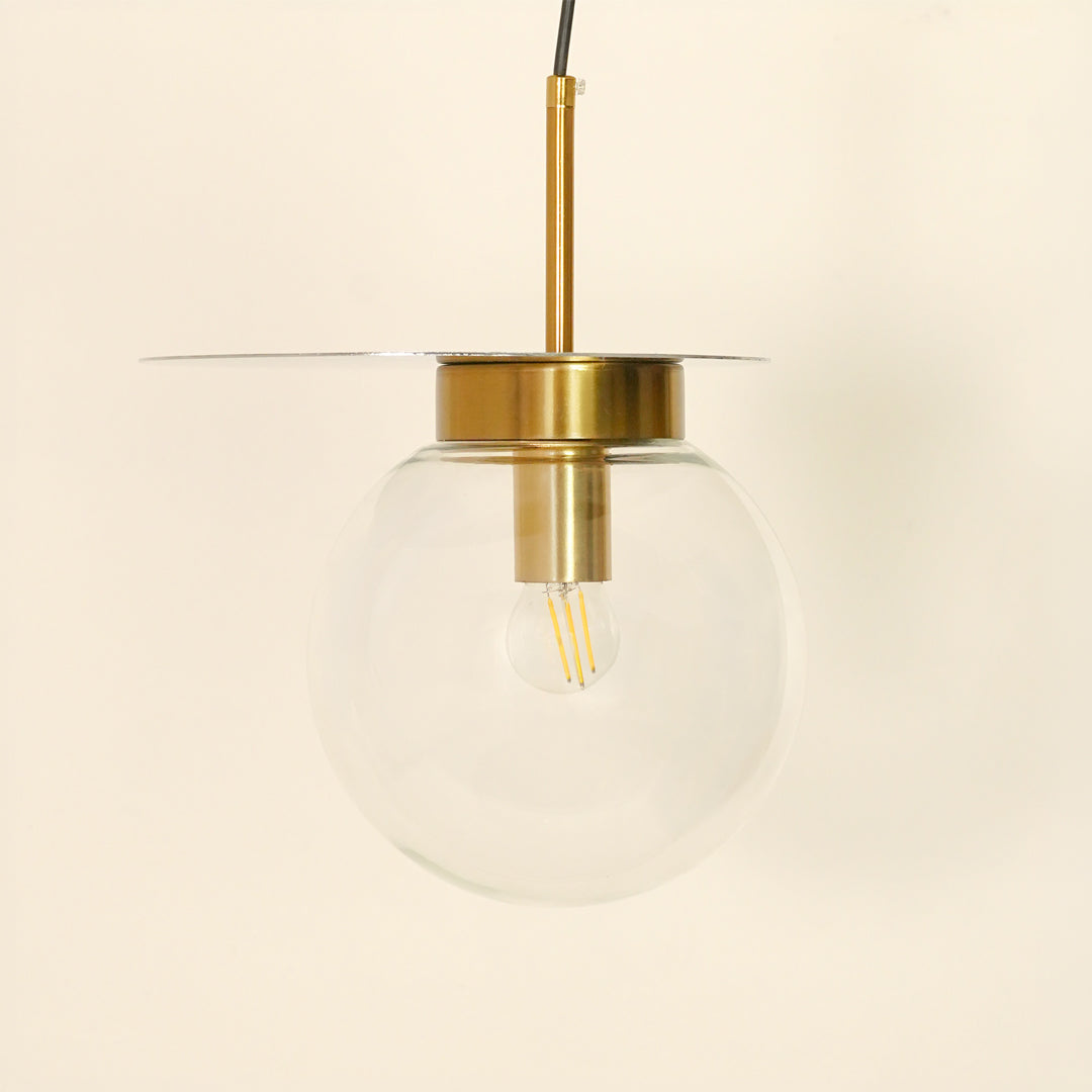 A modern pendant light with a clear glass globe shade, a brass-finished metal canopy and rod, and a thin horizontal glass or metal disc shade, shown against a plain cream background.
