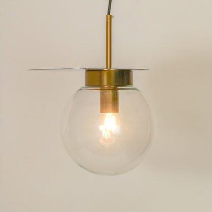 A modern pendant light with a clear glass globe shade, a brass-finished central cap and rod, and a thin horizontal glass disc shade, shown against a plain light beige background.